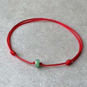 May include: A red string bracelet with a green jade bead. The bracelet is tied with a knot at each end.