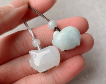 May include: Two jade pig pendants on a brown and white cord. The pendants are carved in the shape of pigs and are a light green and white color.