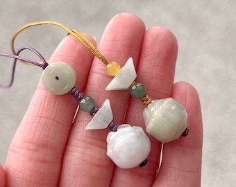 May include: Two jade stone charms on a purple and gold cord. The charms are a round disc with a hole in the center and a carved flower shape.