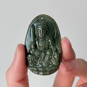 以下が含まれることがあります： 剣を持った座った人物が彫刻された濃い緑色の翡翠のペンダント。人物は、より小さな人物と複雑なディテールに囲まれています。
