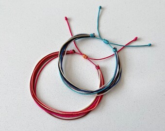 Può includere: Due braccialetti in corda regolabili. Un braccialetto presenta diversi fili sottili nelle tonalità del rosso, rosa e bianco con un nodo scorsoio rosa. Il secondo braccialetto è composto da fili sottili blu, bianchi e neri con un nodo scorsoio turchese.