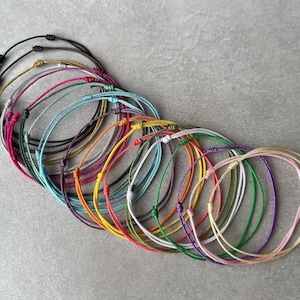 May include: A selection of colourful string bracelets in shades like black, blue, green, yellow, orange, pink, and purple. Each bracelet has a simple knot closure. The bracelets are displayed on a grey surface.