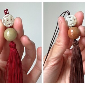 Puede incluir: Un borla decorativa con una cuenta blanca tallada, una cuenta redonda verde y una borla roja. La borla es sostenida por una mano sobre un fondo blanco. La segunda imagen muestra una borla similar con una borla marr&oacute;n.