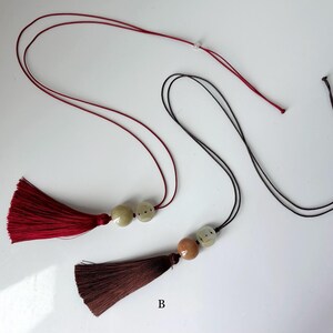 Puede incluir: Dos collares con borlas. Uno tiene una borla burdeos con cord&oacute;n a juego y dos cuentas blanquecinas. El otro tiene una borla marr&oacute;n con un cord&oacute;n negro y dos cuentas, una marr&oacute;n y otra blanquecina. Ambos tienen un cierre blanco.