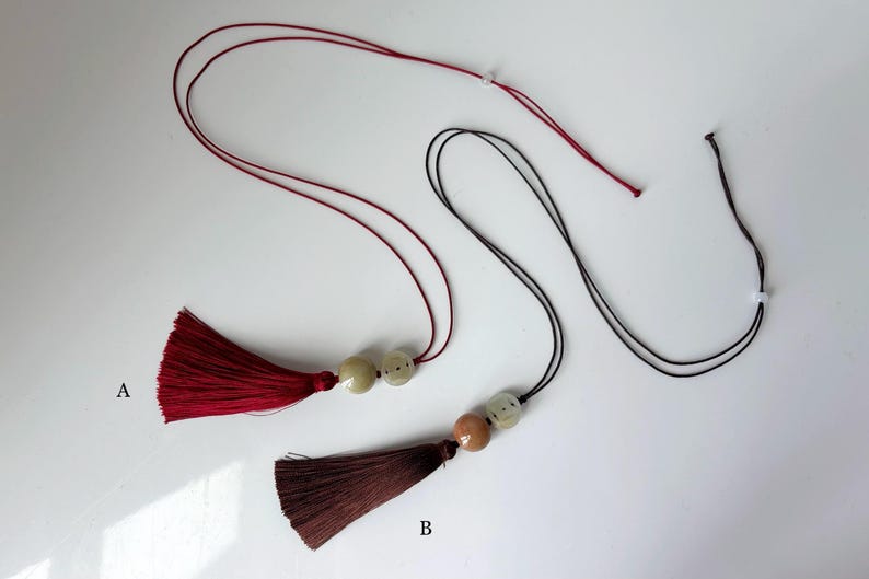 Puede incluir: Dos collares con borlas. Uno tiene una borla burdeos con cord&oacute;n a juego y dos cuentas blanquecinas. El otro tiene una borla marr&oacute;n con un cord&oacute;n negro y dos cuentas, una marr&oacute;n y otra blanquecina. Ambos tienen un cierre blanco.