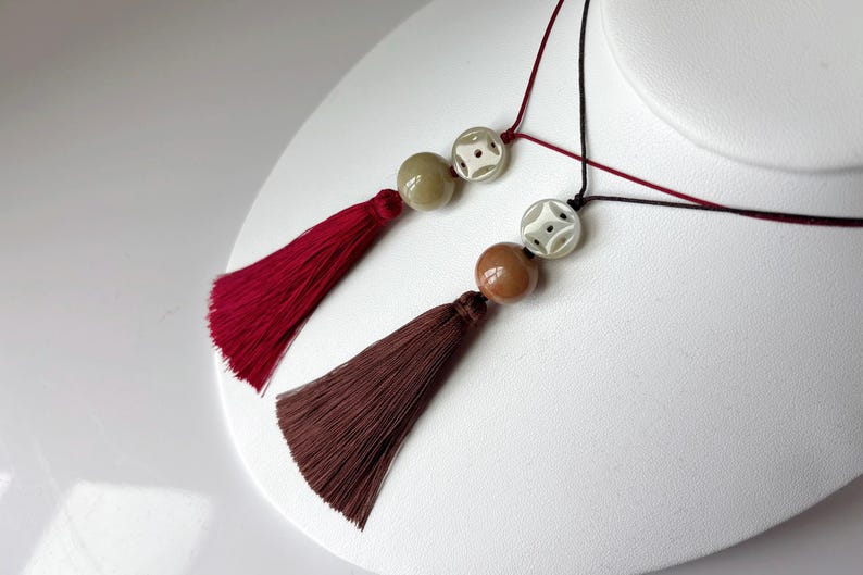 Puede incluir: Dos collares con borlas y cuentas. Uno tiene una borla burdeos y una cuenta verde claro, el otro tiene una borla marr&oacute;n y una cuenta marr&oacute;n. Ambos collares tienen una cuenta blanca con un dise&ntilde;o geom&eacute;trico.