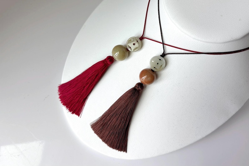 Puede incluir: Dos collares con borlas exhibidos sobre una superficie blanca. Uno presenta una borla burdeos con una cuenta beige y estampada. El otro tiene una borla marr&oacute;n con una cuenta marr&oacute;n y estampada. Ambos tienen cordones negros y rojos.
