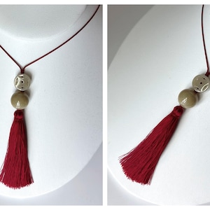 Puede incluir: Un collar con borlas burdeos con un cord&oacute;n rojo. El collar presenta dos cuentas redondas, una con un dise&ntilde;o estampado, y una cuenta m&aacute;s grande de color s&oacute;lido. La borla es larga y completa, a&ntilde;adiendo un toque dram&aacute;tico a la joya.