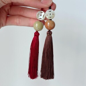 Puede incluir: Dos borlas decorativas, cada una con un dise&ntilde;o diferente. Una presenta una borla roja con una cuenta verde y una cuenta blanca tallada. La otra tiene una borla marr&oacute;n con una cuenta marr&oacute;n y una cuenta blanca tallada.