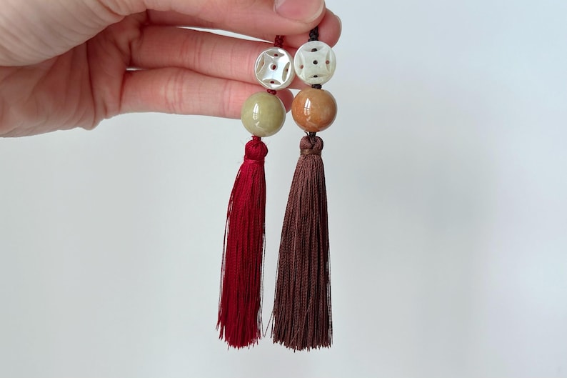 Puede incluir: Dos borlas decorativas, cada una con un dise&ntilde;o diferente. Una presenta una borla roja con una cuenta verde y una cuenta blanca tallada. La otra tiene una borla marr&oacute;n con una cuenta marr&oacute;n y una cuenta blanca tallada.