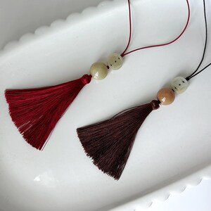 Puede incluir: Dos borlas decorativas con cuentas. Una borla es de color burdeos intenso y la otra marr&oacute;n oscuro. Ambas tienen una textura sedosa y con flecos y est&aacute;n suspendidas por finos cordones. Las borlas descansan sobre un plato blanco con borde festoneado.
