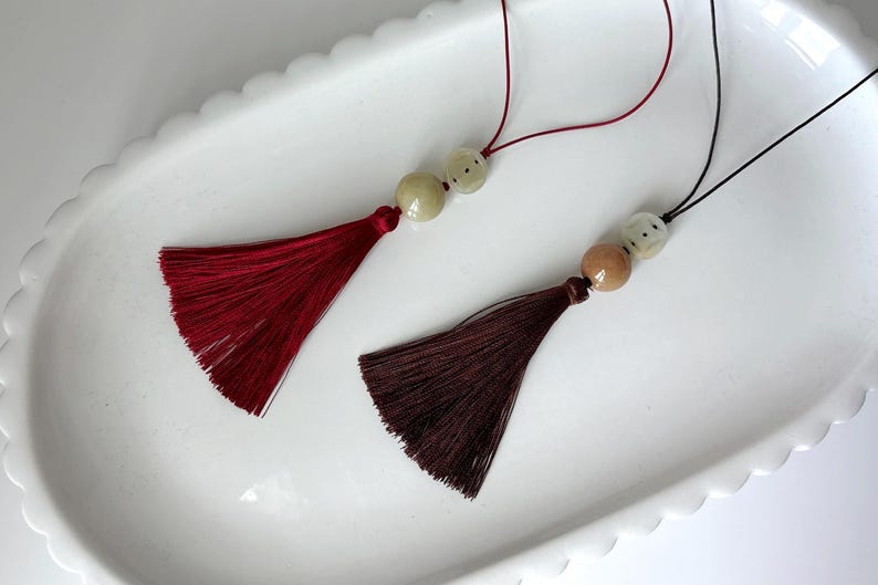 Puede incluir: Dos borlas decorativas con cuentas. Una borla es de color burdeos intenso y la otra marr&oacute;n oscuro. Ambas tienen una textura sedosa y con flecos y est&aacute;n suspendidas por finos cordones. Las borlas descansan sobre un plato blanco con borde festoneado.
