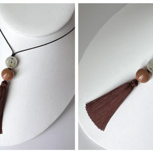 Puede incluir: Un collar con un cord&oacute;n negro, con una cuenta clara estampada, una cuenta marr&oacute;n y una borla marr&oacute;n. El collar se exhibe en un busto de maniqu&iacute; blanco, mostrando su dise&ntilde;o y longitud.
