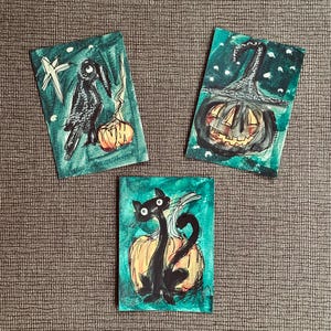 May include: Three rectangular art prints featuring hand-drawn illustrations on a teal background. One shows a black bird with a pumpkin, another a jack-o'-lantern wearing a hat, and the third a black cat sitting in front of a large orange pumpkin.