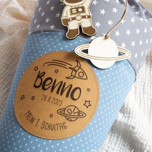 Könnte beinhalten: Ein blauer und grauer Stoffkegel mit weißen Sternen, einem Holz-Astronauten, einem Holz-Planeten mit Ringen und einem braunen Etikett mit der Aufschrift "Benno 28.8.2023 Mein 1. Schultag".