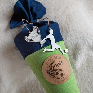 Könnte beinhalten: Ein grüner und blauer Schultüte mit einem Fußball und einer Trophäe. Die Tüte hat einen braunen Kreis mit dem Text "Ich bin in 1. Klasse!" und "Jannis" mit einer Fußballgrafik. Das Datum "9. September 2024" ist ebenfalls auf dem Kreis.