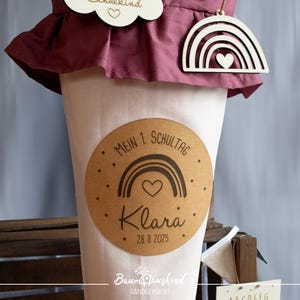 Könnte beinhalten: Ein brauner Papierkegel mit einem weißen Etikett, auf dem "Mein 1. Schultag" mit einem Regenbogen und einem Herzdesign steht. Das Etikett enthält auch den Namen "Klara" und das Datum "28.8.2025".