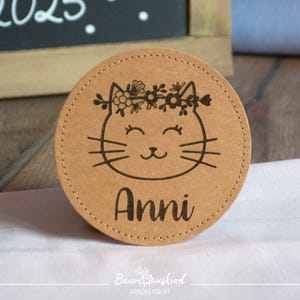 Könnte beinhalten: Ein runder, brauner Holz-Untersetzer mit einem Katzenkopf-Design. Die Katze trägt eine Blumenkrone und der Name "Anni" ist unter der Katze eingraviert.