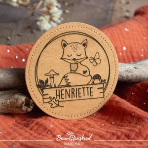 Könnte beinhalten: Ein runder, brauner Lederuntersetzer mit einem Fuchsdesign, Pilzen, einem Schmetterling und dem Namen "Henriette" eingraviert. Der Untersetzer ist handgefertigt und hat ein rustikales, natürliches Aussehen.