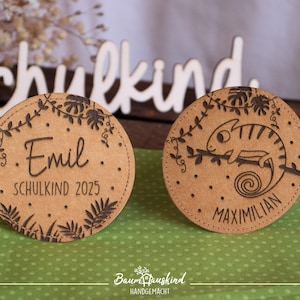 Könnte beinhalten: Zwei runde Holz-Namensschilder mit Dschungel-Motiv. Das Schild links zeigt "Emil Schulkind 2025" und das Schild rechts zeigt "Maximilian" mit einer Chamäleon-Illustration.