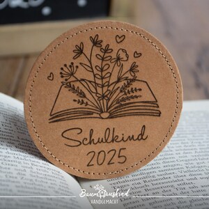 Könnte beinhalten: Ein brauner Lederuntersetzer mit einem Design eines offenen Buches mit Blumen und dem Text "Schulkind 2025".