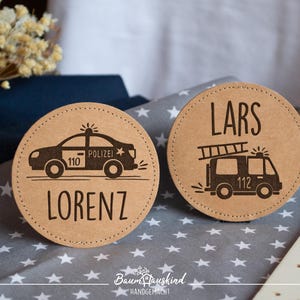 Könnte beinhalten: Zwei runde Holz-Platzkarte mit den Namen "Lars" und "Lorenz" darauf gedruckt. Die Karte mit "Lars" hat ein Feuerwehrauto mit der Nummer 112 darauf. Die Karte mit "Lorenz" hat ein Polizeiauto mit der Nummer 110 darauf.