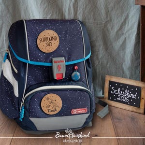 Könnte beinhalten: Ein blauer und schwarzer Rucksack mit Sternenmuster. Er hat ein Holzschild mit der Aufschrift "Schulkind 2023" und ein weiteres Schild mit der Aufschrift "Noah".