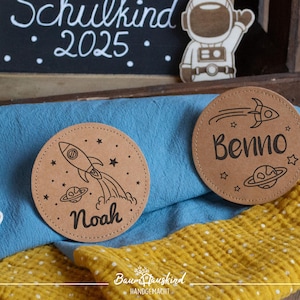 Könnte beinhalten: Zwei braune, runde, hölzerne Namensschilder mit Weltraumthema. Ein Schild hat eine Rakete und den Namen "Noah" und das andere Schild hat einen Kometen und den Namen "Benno".