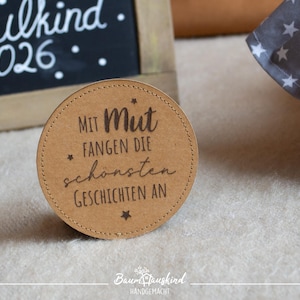 Könnte beinhalten: Ein runder, hellbrauner Untersetzer mit dem Text "Mit Mut fangen die schönsten Geschichten an" in Braun. Der Untersetzer hat einen genähten Rand und steht vor einem weichen, hellfarbigen Hintergrund. Im Hintergrund befindet sich eine Tafel mit dem Text "Schulkind 2026".