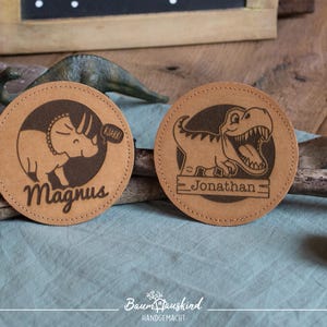 Könnte beinhalten: Zwei braune Lederuntersetzer mit Dinosauriermotiven und personalisierten Namen. Der Untersetzer links zeigt einen Triceratops mit dem Namen "Magnus" und der Untersetzer rechts einen T-Rex mit dem Namen "Jonathan".