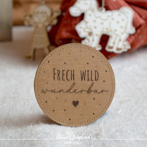 Könnte beinhalten: Ein runder, hellbrauner Untersetzer mit den Worten "FRECH WILD wunderbar" und einem kleinen Herzen, das in die Oberfläche geätzt ist. Der Untersetzer hat einen gepunkteten Rand. Holzdekorationen im Hintergrund.