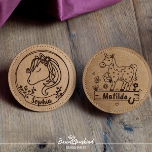 Könnte beinhalten: Zwei runde, braune Lederuntersetzer mit gravierten Designs. Ein Untersetzer zeigt ein Einhorn mit dem Namen "Sophia" und der andere ein Pferd mit dem Namen "Matilda".