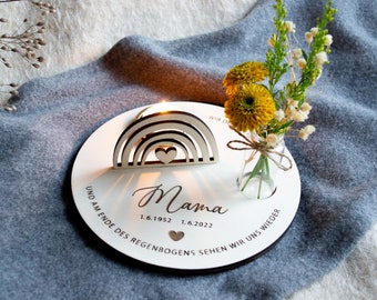 Puede incluir: Una placa conmemorativa de madera con un diseño de arcoíris y la inscripción "Mama". Las fechas "1.6.1952" y "1.6.2022" también están inscritas en la placa. Un pequeño jarrón con flores amarillas se coloca en la placa. La placa está sobre una superficie gris.