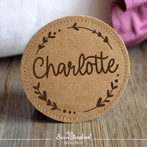 Könnte beinhalten: Ein rundes, braunes Leder-Etikett mit einem genähten Rand. Das Etikett hat ein florales Design und den Namen "Charlotte" in einer kursiv geschriebenen Schrift.