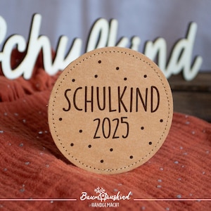 Könnte beinhalten: Ein rundes, braunes Holzschild mit dem Text "SCHULKIND 2025" in schwarzer Schrift. Das Schild ist mit kleinen weißen Punkten verziert.