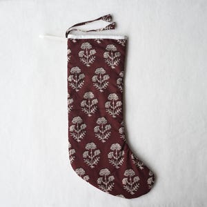 Könnte beinhalten: Ein tiefroter Weihnachtsstrumpf mit einem sich wiederholenden weißen Blumenmuster im Blockdruck. Der Strumpf hat einen weißen Stoffsaum an der oberen Öffnung und eine dünne weiße Stoffschlaufe an der Ecke zum Aufhängen des Artikels.