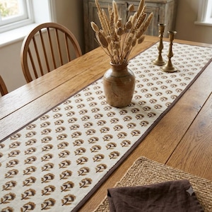 Può includere: Un lungo runner da tavola rettangolare con un motivo floreale stampato a blocchi marrone ripetuto su una base in tessuto color bianco sporco con un bordo marrone scuro, centrato su un tavolo in legno con un vaso in ceramica di erbe essiccate.