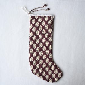 Op de afbeelding: Een kerstkous met een diepe kastanjebruine achtergrond en een herhalend crèmekleurig bloemenpatroon in blokdruk. De kous heeft een witte stoffen boord aan de bovenkant en een bijpassende stoffen lus om op te hangen aan de linkerkant.