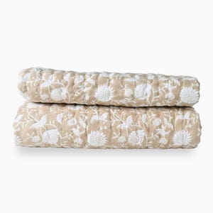 Puede incluir: Dos edredones beige con estampados florales blancos apilados. Los edredones tienen un diseño acolchado y una paleta de colores neutros, adecuados para un dormitorio o sala de estar.