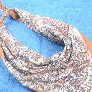 Op de afbeelding: Een paisley bandana in crème, bruin en blauw. De bandana is gevouwen en geknoopt, met een effen bruine rand. De stof heeft een gedetailleerd bloemmotief.