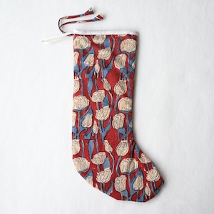 Op de afbeelding: Een kerstkous van rode stof met een herhalend patroon van crèmekleurige tulpachtige bloemen met blauwe stelen en bladeren. De kous heeft een witte boord aan de bovenkant en is voorzien van twee dunne stoffen linten om op te hangen.