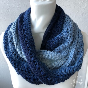 Peut inclure: Une écharpe infinie en crochet dans des tons de bleu, du bleu clair au bleu foncé. L'écharpe est réalisée avec un point texturé et a un aspect délicat et ajouré. L'écharpe est présentée sur un mannequin blanc.