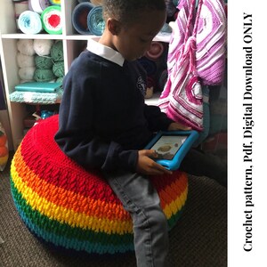 May include: A child sits on a large, colorful rainbow crochet pouf. The pouf is made of thick yarn and has a red, orange, yellow, green, blue, and purple color scheme. The child is using a blue tablet device.
