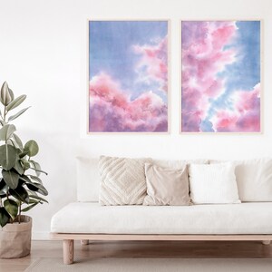 Puede incluir: Dos pinturas de acuarela enmarcadas de nubes rosas y azules. Las pinturas est&aacute;n colgadas en una pared blanca sobre un sof&aacute; blanco con almohadas.