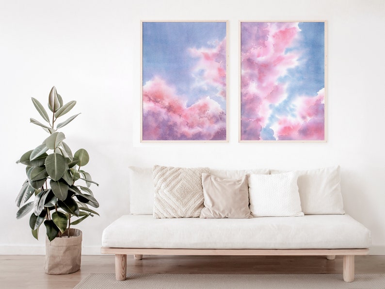Puede incluir: Dos pinturas de acuarela enmarcadas de nubes rosas y azules. Las pinturas est&aacute;n colgadas en una pared blanca sobre un sof&aacute; blanco con almohadas.
