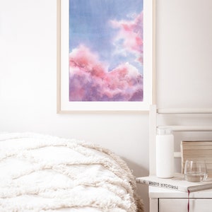Puede incluir: Una pintura de acuarela enmarcada de un cielo nublado rosa y morado. La pintura est&aacute; colgada en una pared blanca sobre una cama blanca con una manta blanca.