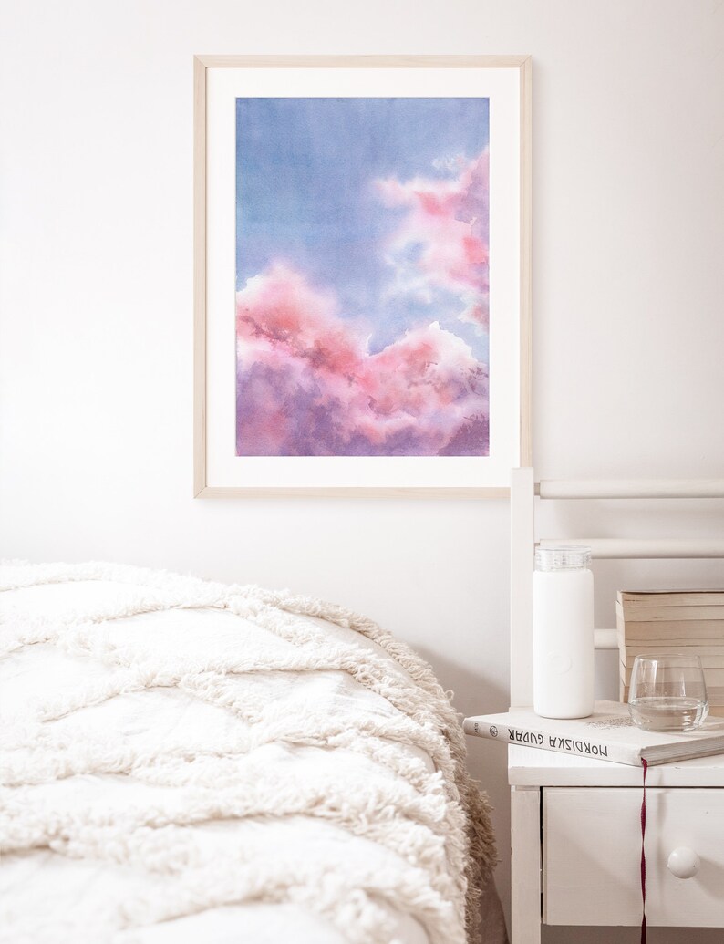 Puede incluir: Una pintura de acuarela enmarcada de un cielo nublado rosa y morado. La pintura est&aacute; colgada en una pared blanca sobre una cama blanca con una manta blanca.
