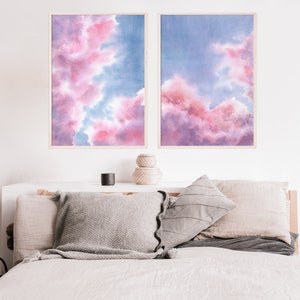 Puede incluir: Dos pinturas de acuarela enmarcadas de nubes rosas y azules. Las pinturas est&aacute;n colgadas en una pared blanca sobre una cama con almohadas grises y beige.