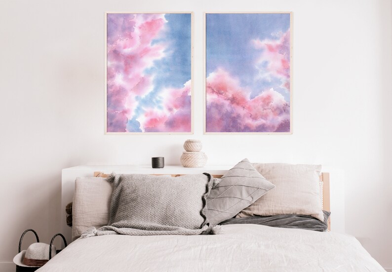 Puede incluir: Dos pinturas de acuarela enmarcadas de nubes rosas y azules. Las pinturas est&aacute;n colgadas en una pared blanca sobre una cama con almohadas grises y beige.