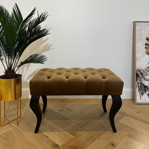 May include: A brown velvet tufted bench with black wooden legs. The bench is sitting on a wood floor with a herringbone pattern.